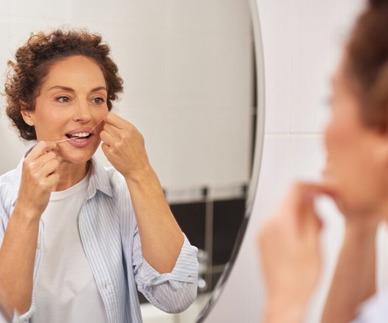 Woman flossing her teeth while looking in a bathroom mirror