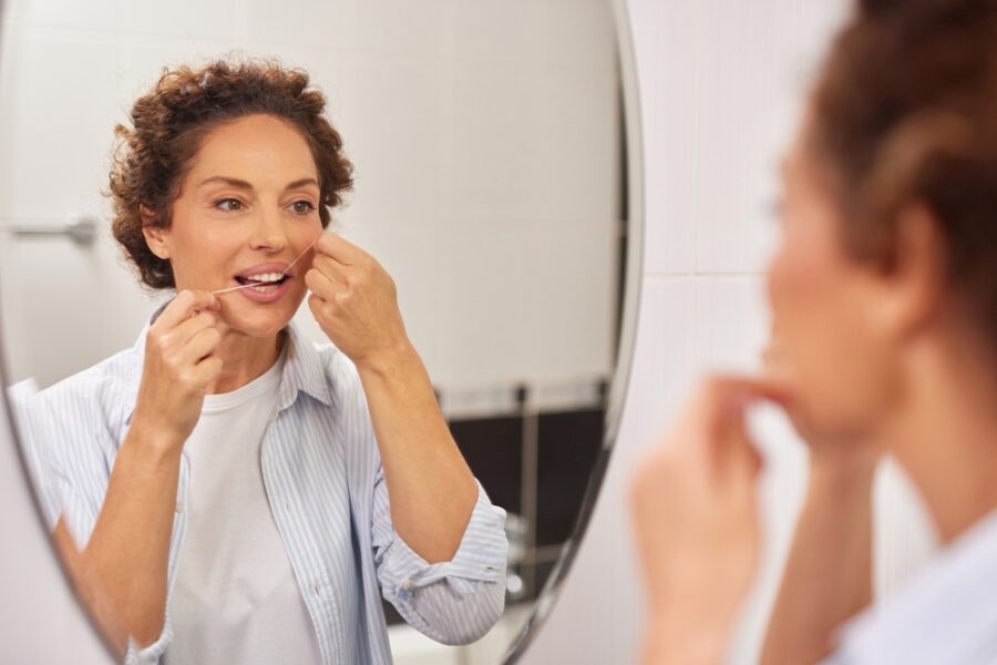 Woman flossing her teeth while looking in a bathroom mirror