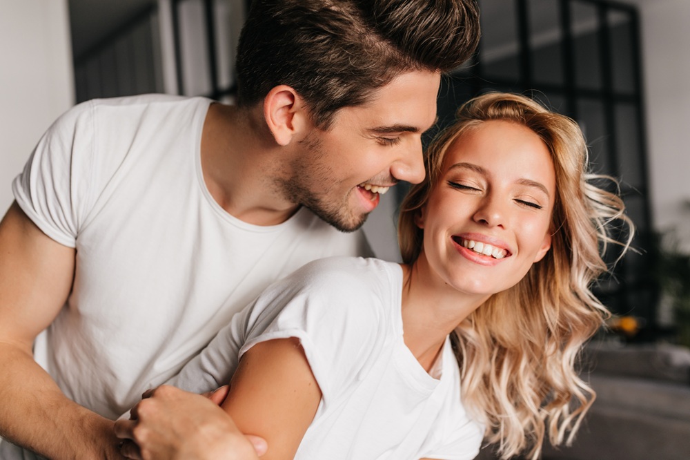 Couple embracing indoors, both smiling with their eyes closed
