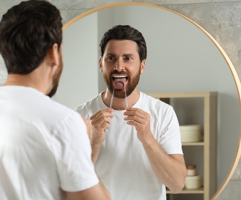 Man using a metal tongue scraper while looking in a round bathroom mirror