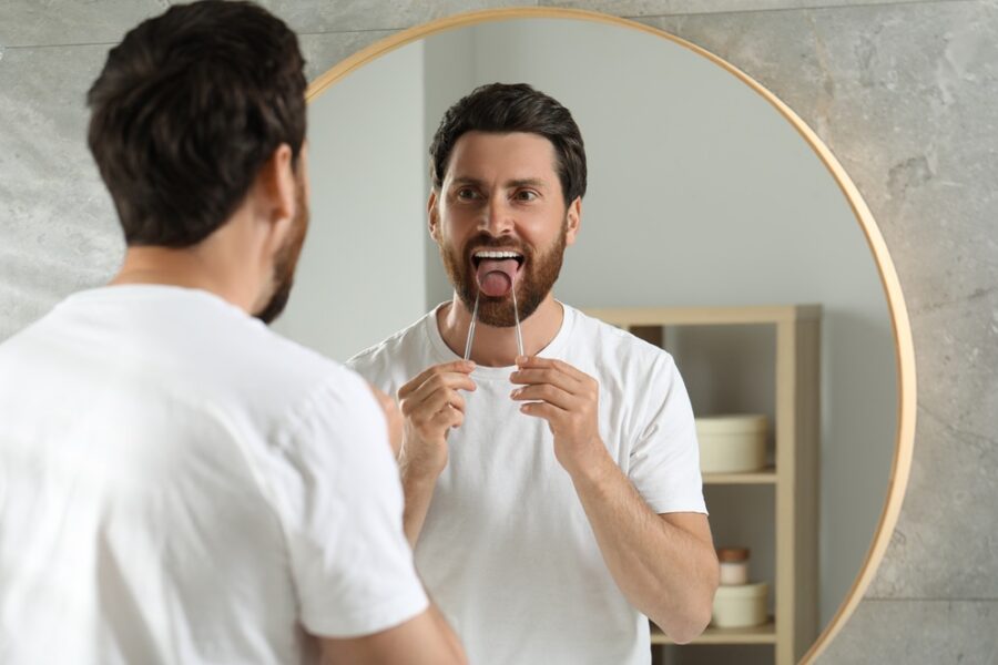 Man using a metal tongue scraper while looking in a round bathroom mirror
