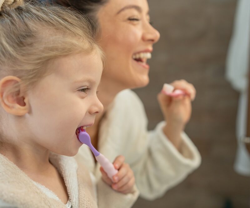 Mother and young daughter smiling while brushing their teeth together as part of a daily oral hygiene routine