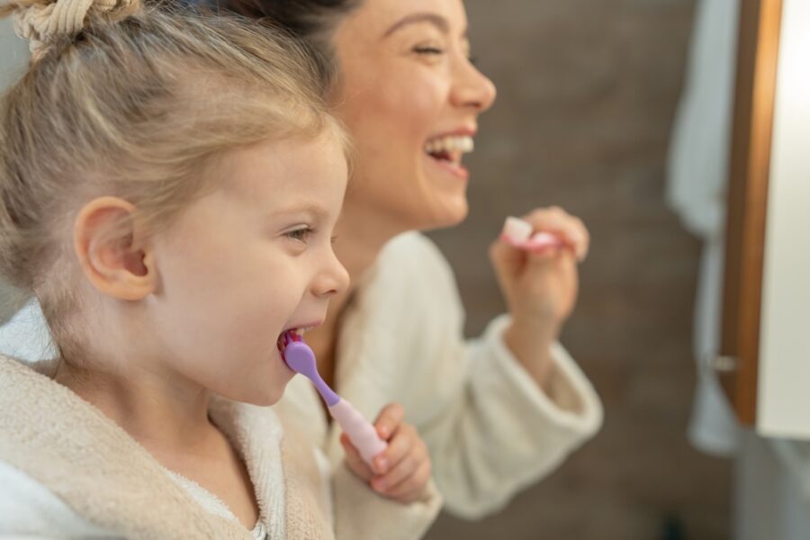 Mother and young daughter smiling while brushing their teeth together as part of a daily oral hygiene routine