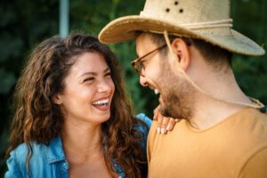 Man and woman laughing face to face outdoors, one wearing a straw hat