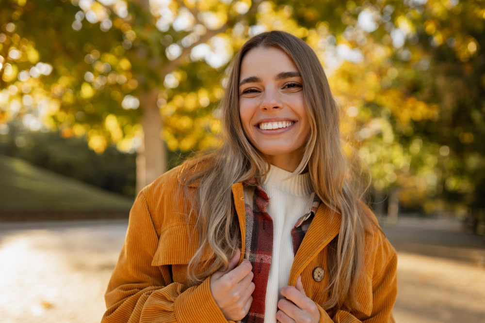 Woman with long wavy hair smiling brightly outdoors in a mustard corduroy jacket during fall