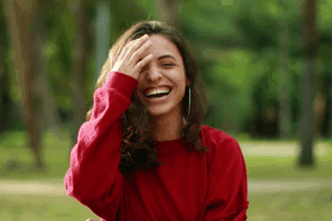 Woman in a red top laughing outdoors with her hand near her cheek